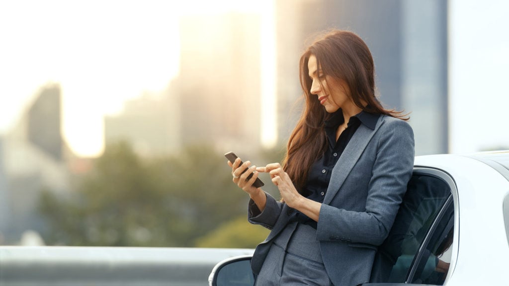 Business Woman Uses Smartphone While Leaning on Her Premium Class Car. Big City with Skyscrapers in the Background.