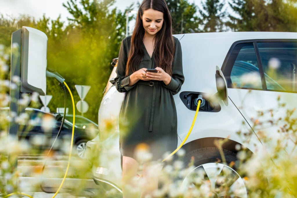 Woman using mobile phone while waiting for electric car to charge in the parking lot